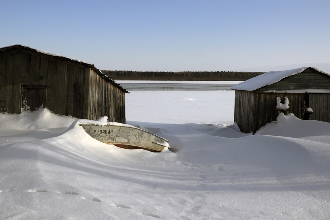 En liten eka delvis täckt av snö, vinterförvarad nära ett båthus vid en frusen sjö.